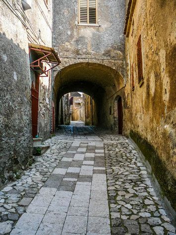 Stretto vicolo lastricato di ciottoli con arco in pietra ed edifici consumati dal tempo che si perdono in lontananza.