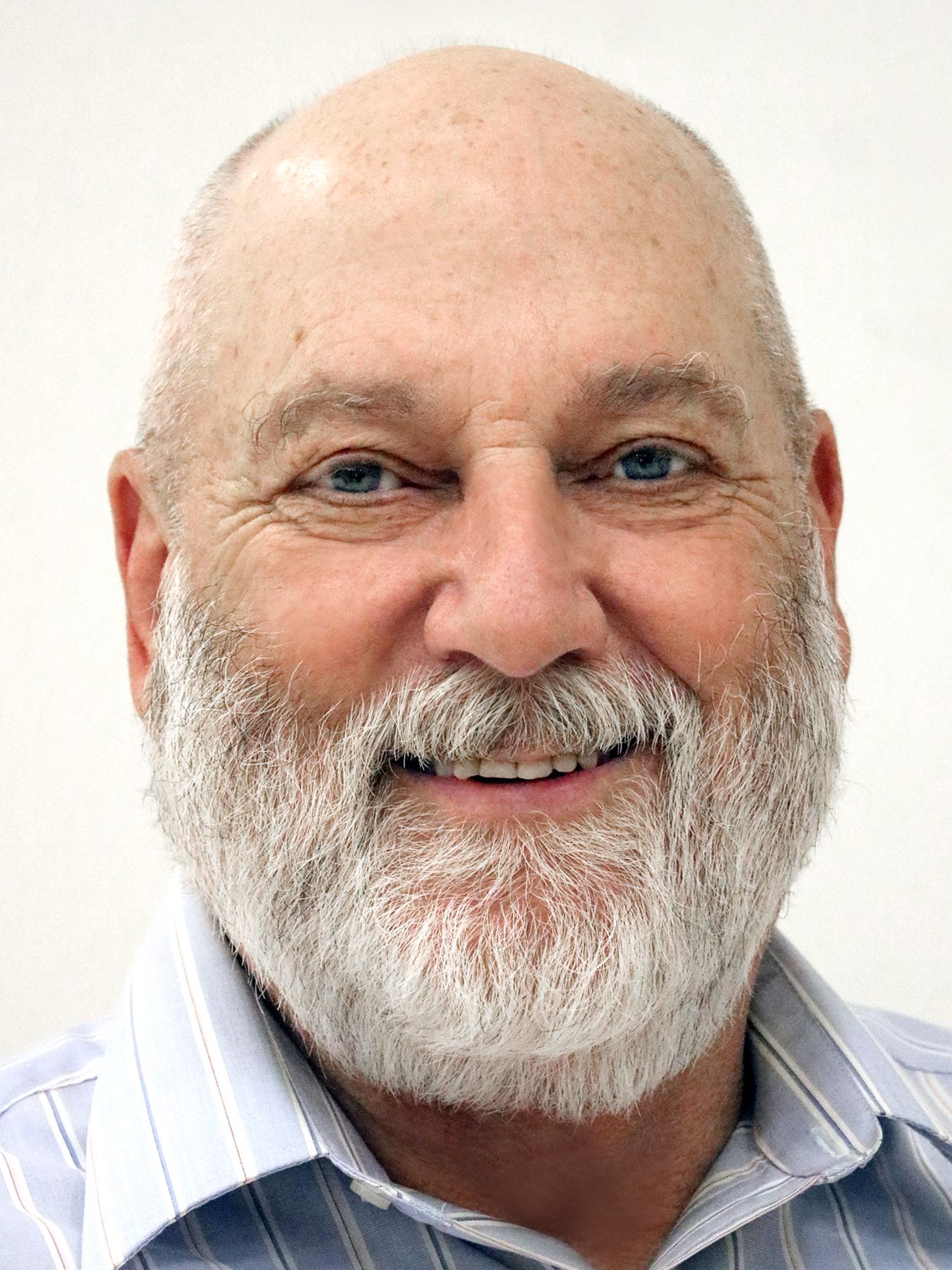 Smiling bald man with a white beard wearing a light blue striped shirt against a white background