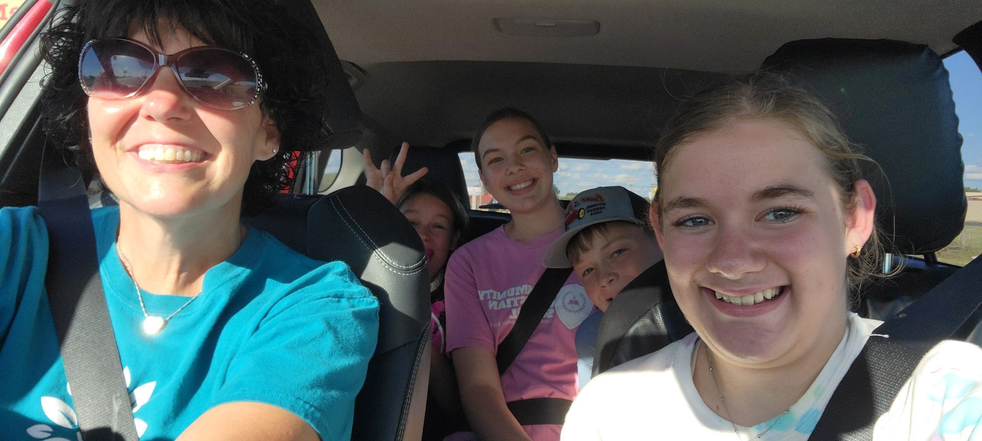 Four smiling people sit in a car, taking a selfie on a sunny day.