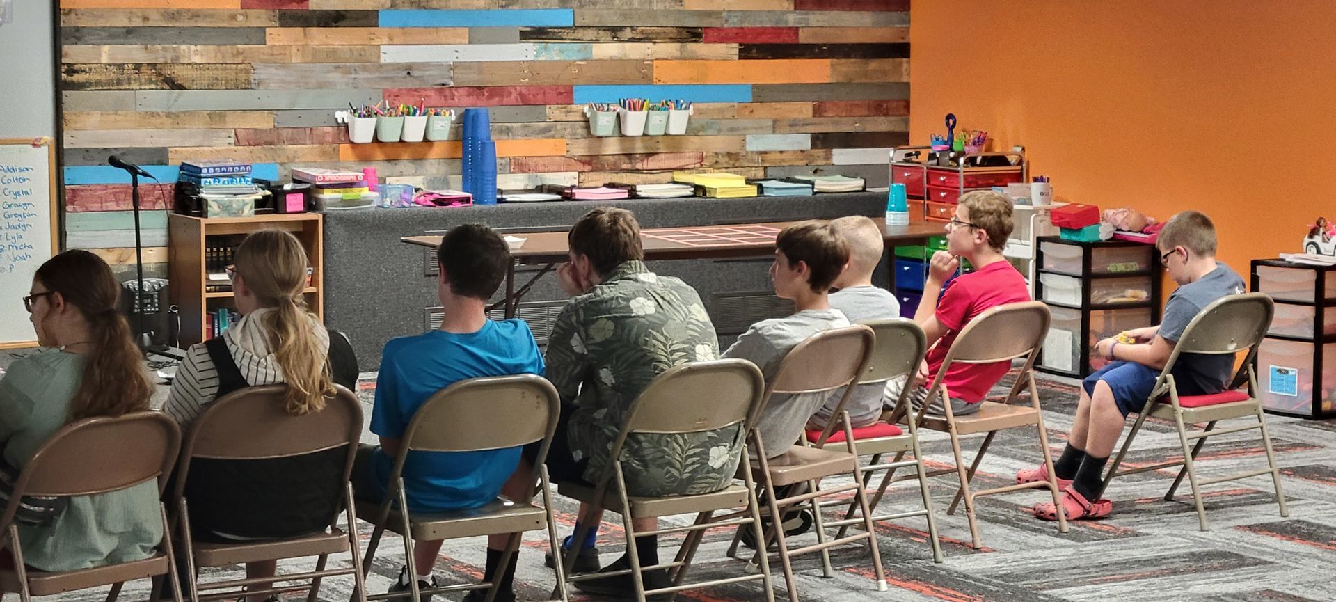 Children seated on folding chairs watching a presentation in a colorful classroom or library.
