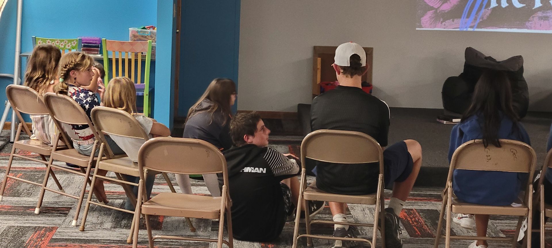 People seated on folding chairs watching a presentation on a large screen indoors.