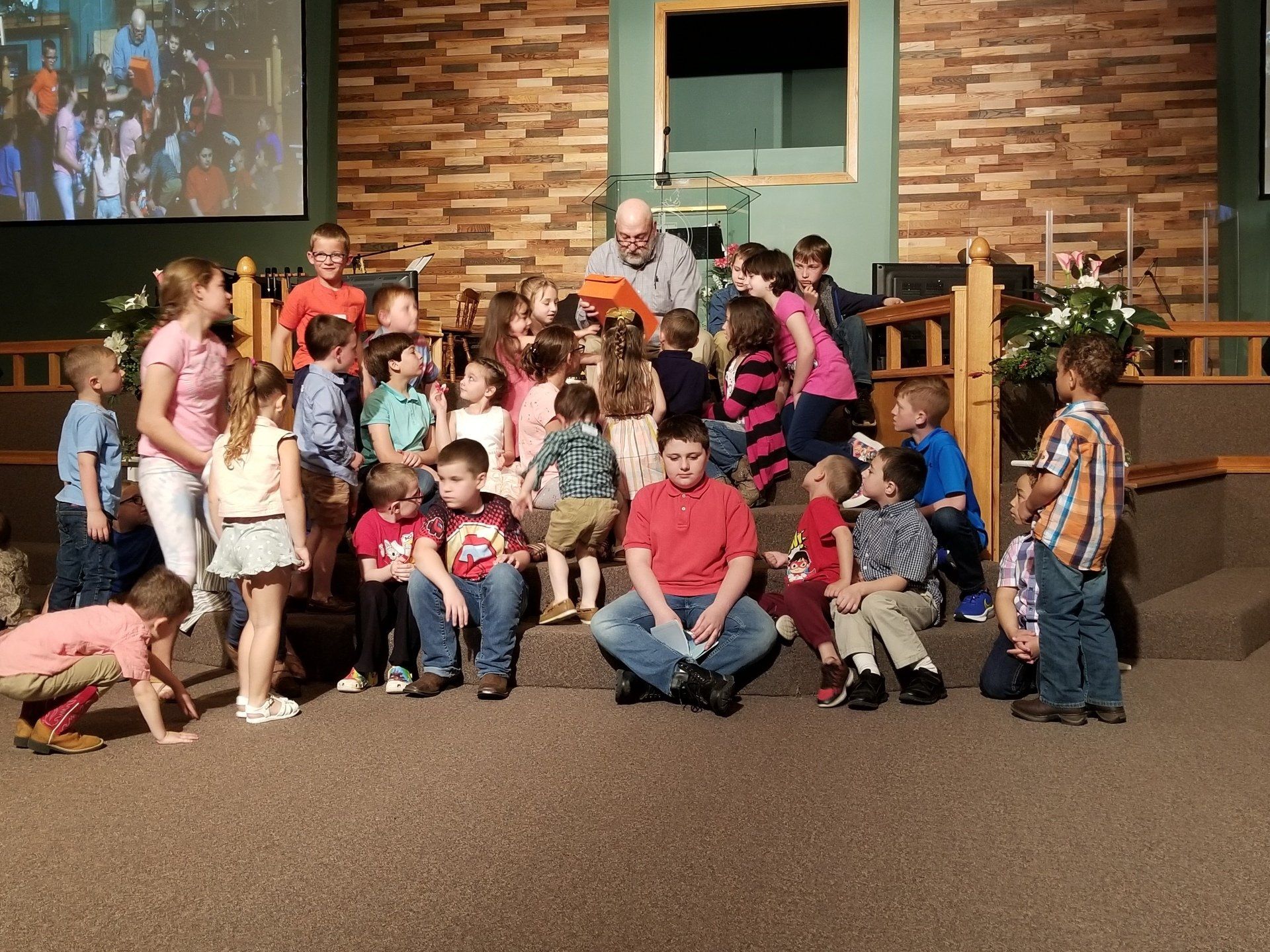 Group of children and adults posing on a church stage in front of a brick wall and cross.