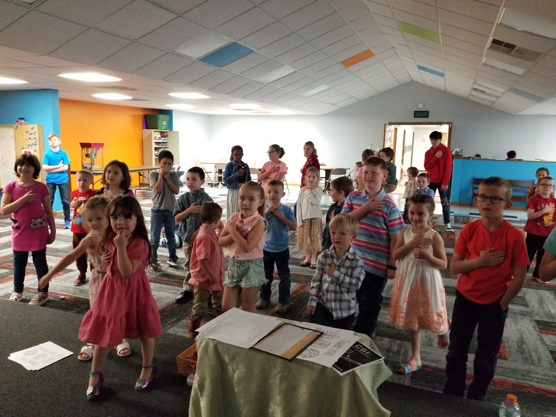 Children dancing in a classroom, with papers on a table in the foreground and bright walls behind them.