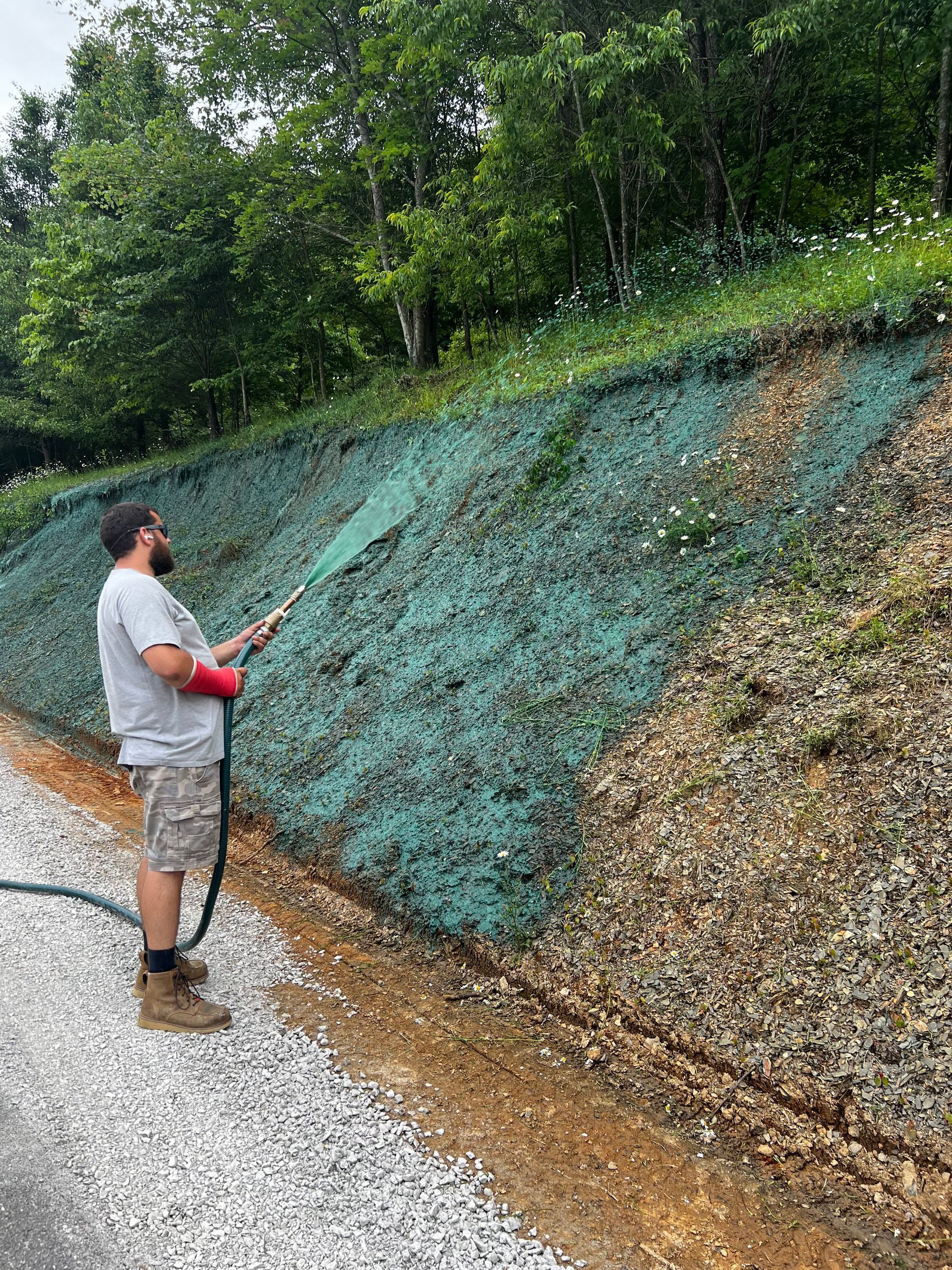 Person standing on a gravel path, spraying a green-covered retaining wall on a wooded hillside.