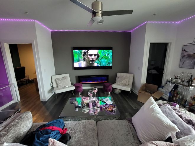 Modern living room with purple LED lighting, wall-mounted TV, sectional sofa, chairs, and coffee table.