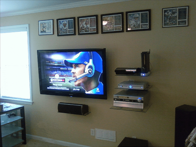 Living room wall with a mounted TV showing a baseball game, framed photos above, and shelves with electronics
