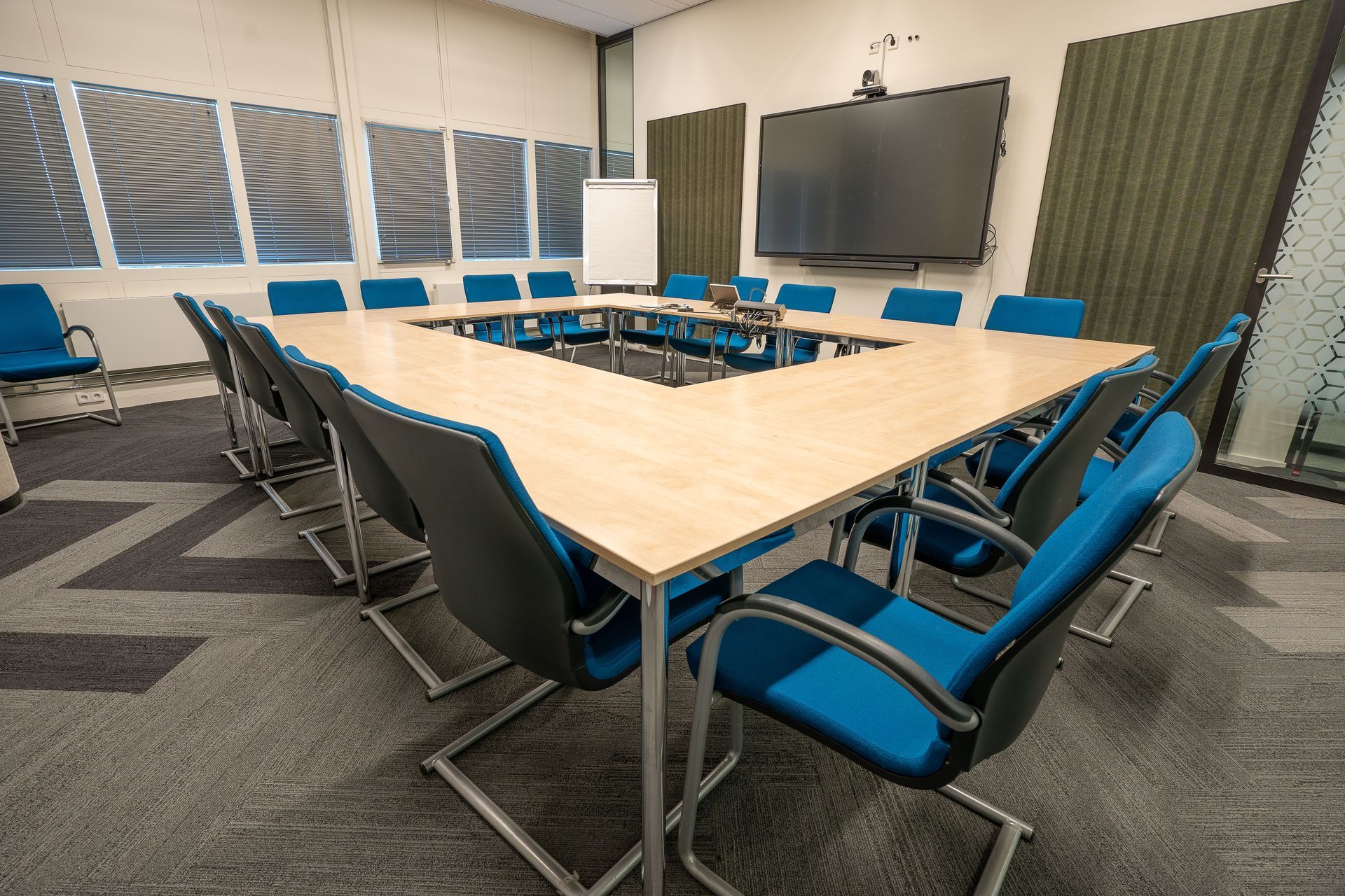 Conference room with blue chairs around a large wood table, wall-mounted TV, and windows with blinds