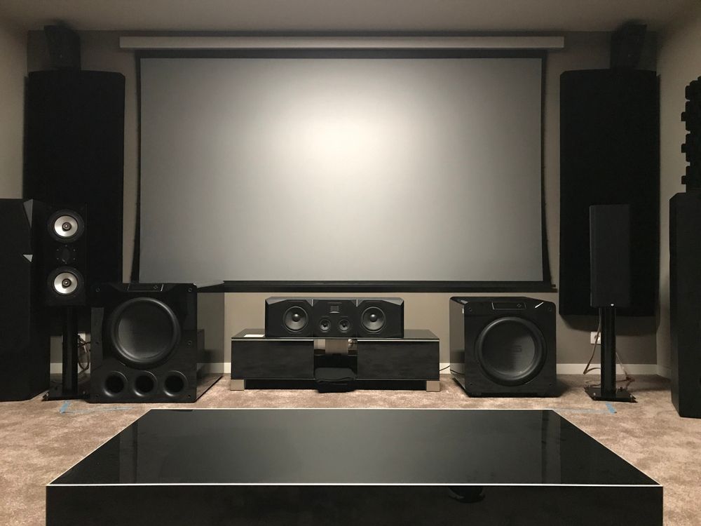 Home theater room with a projector screen, large speakers, subwoofers, and a central AV setup on a carpeted floor