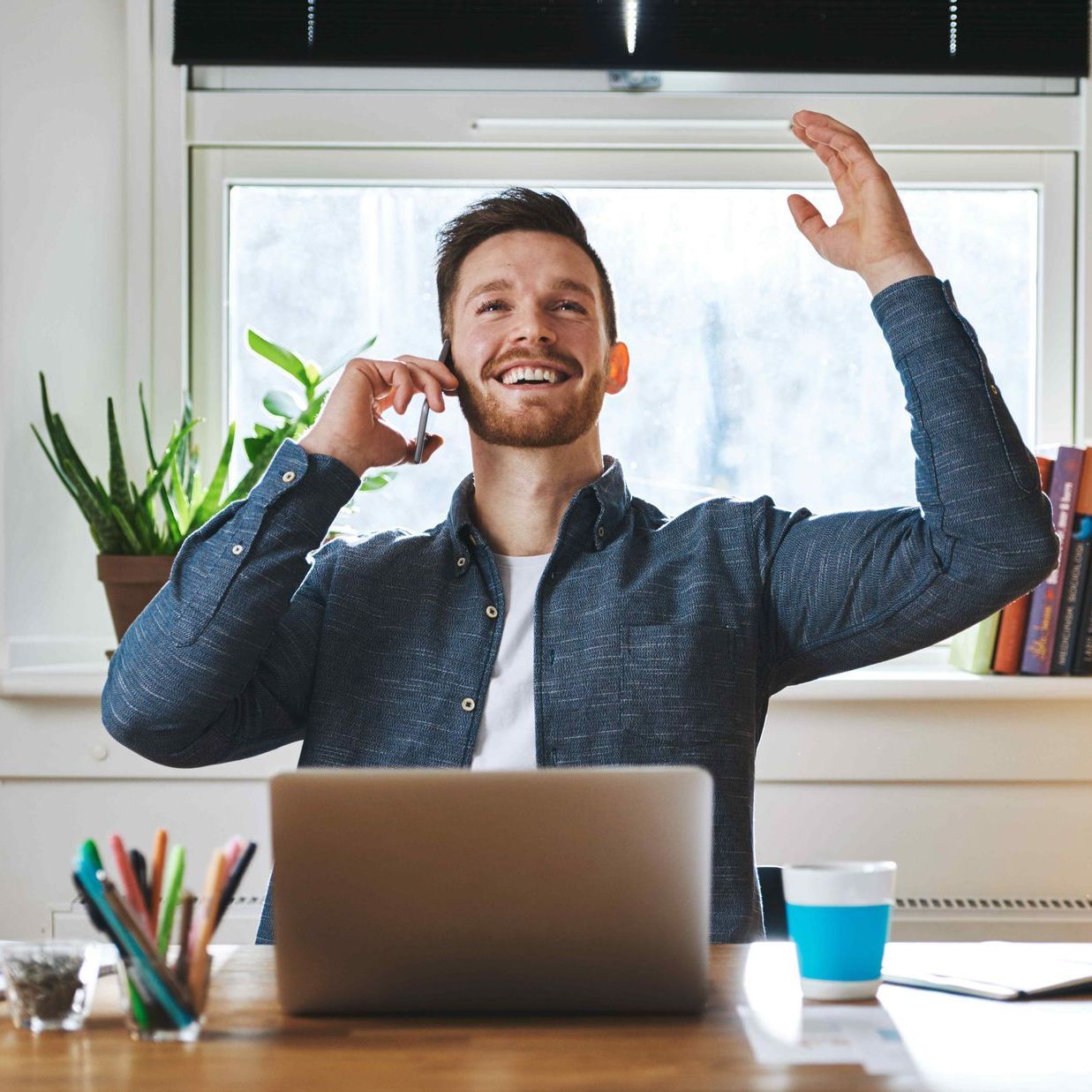 a happy business owner sits in front of his laptop with a phone in one hand and the other hand up in celebration