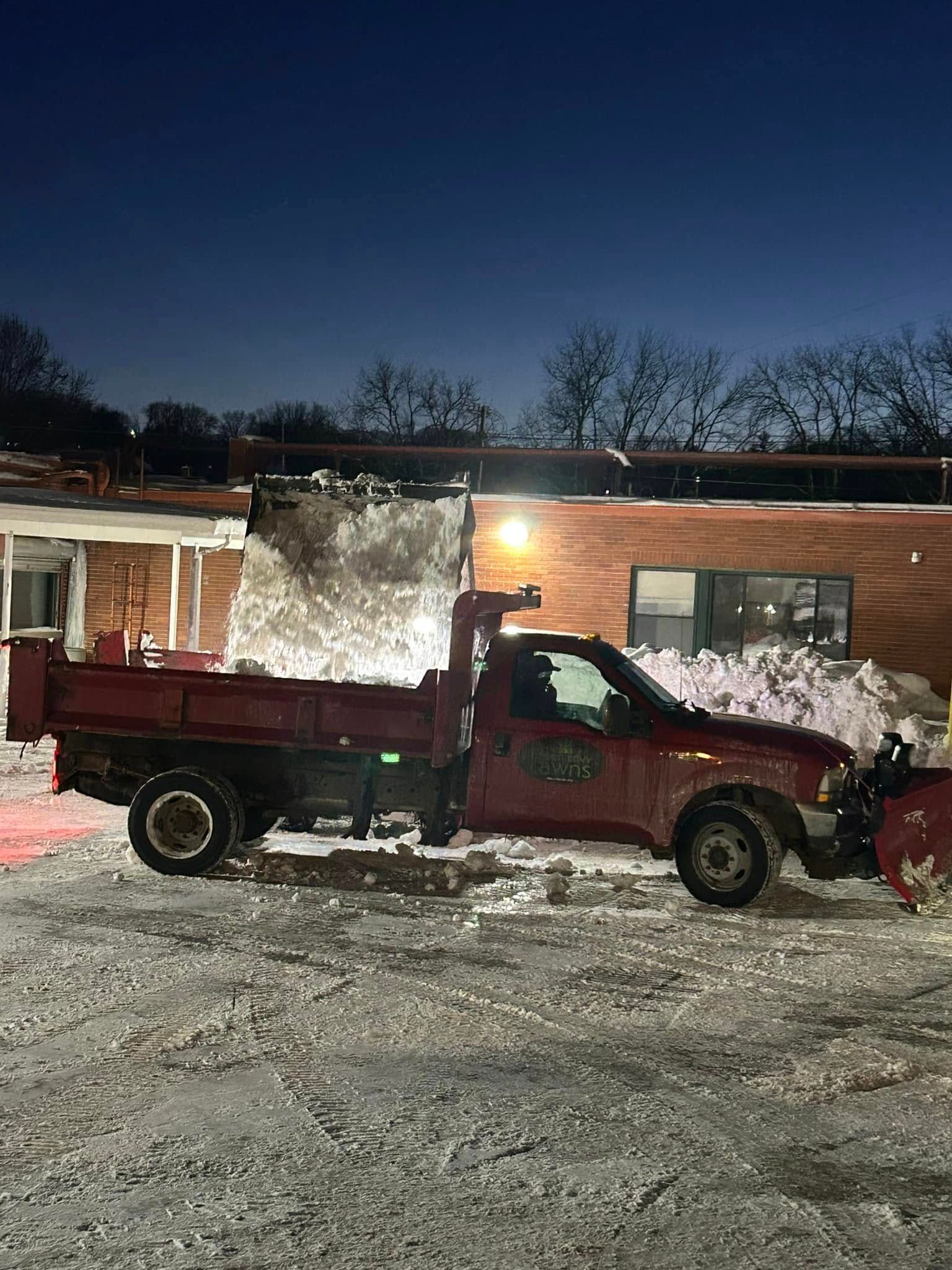 A red dump truck is plowing snow in front of a building at night.