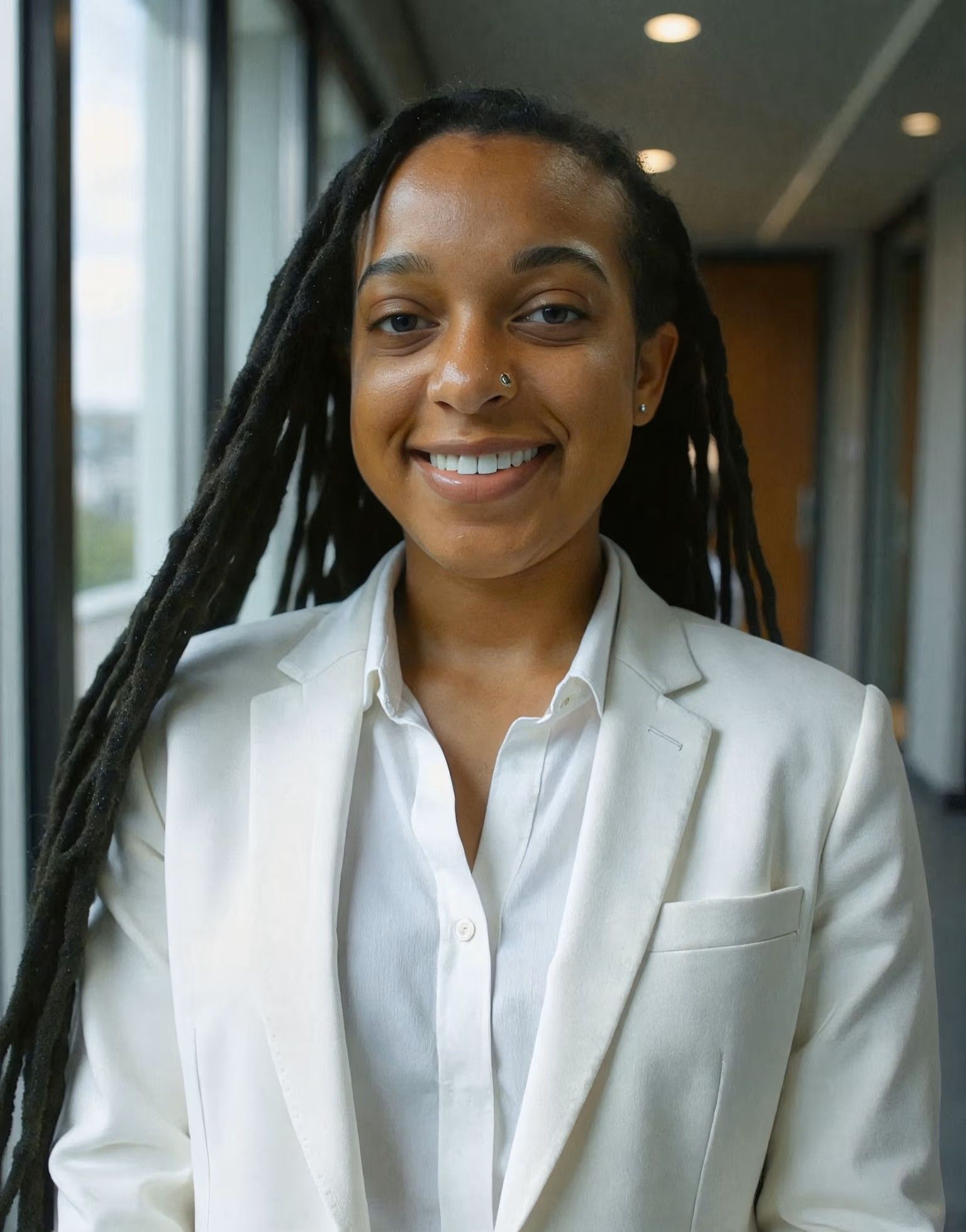 Person in a white suit, smiling, with dreadlocks, in a building with large windows.