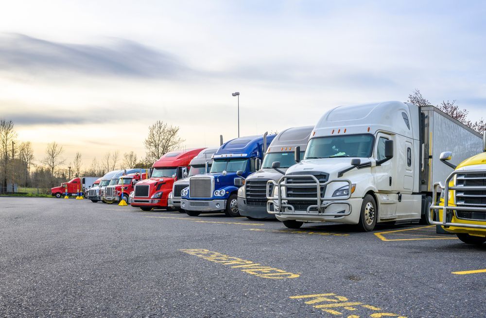 Row of parked semi trucks in a lot, including white, blue, red, and yellow cabs at dusk
