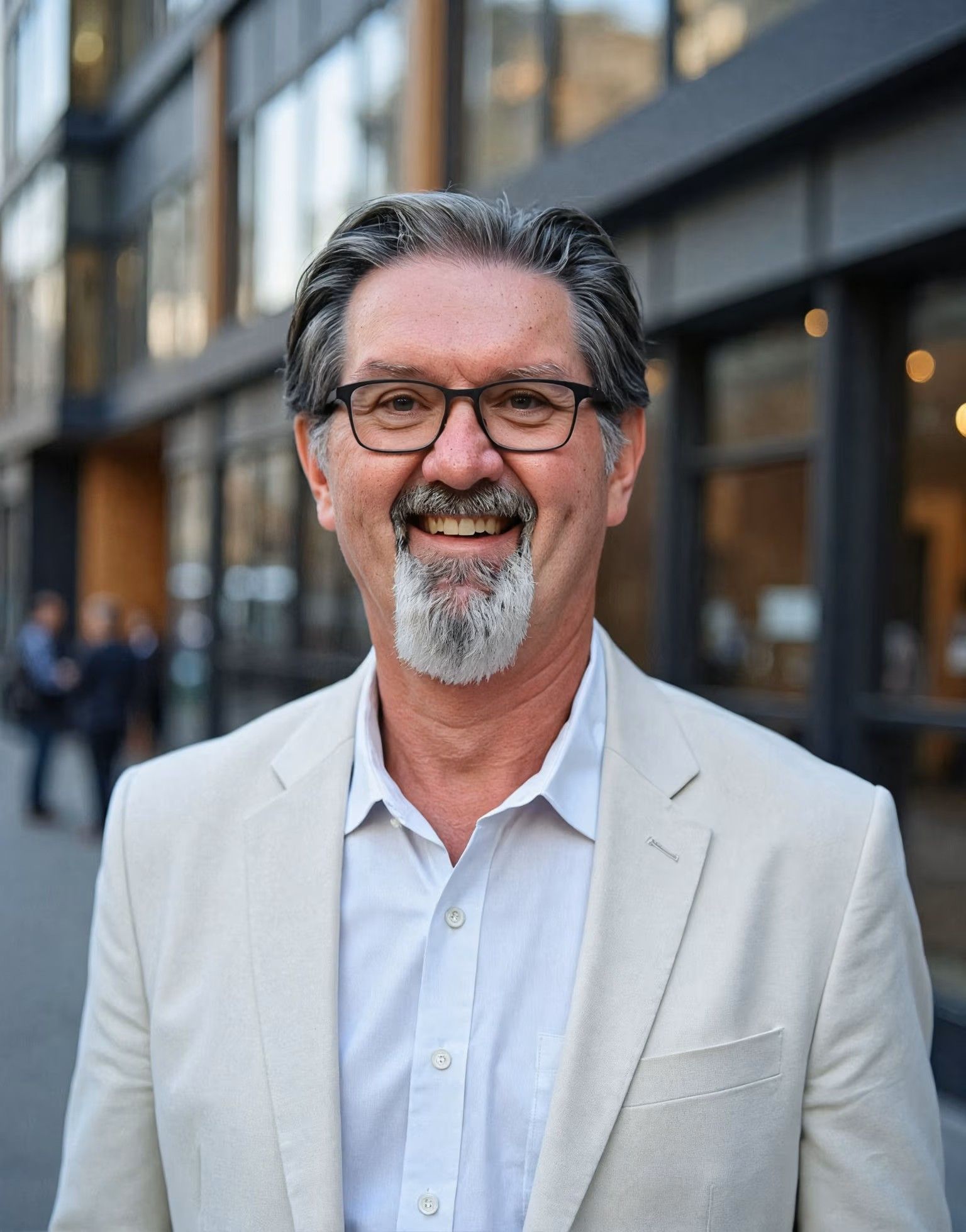 Man with glasses and goatee smiling outdoors, wearing a light-colored jacket.