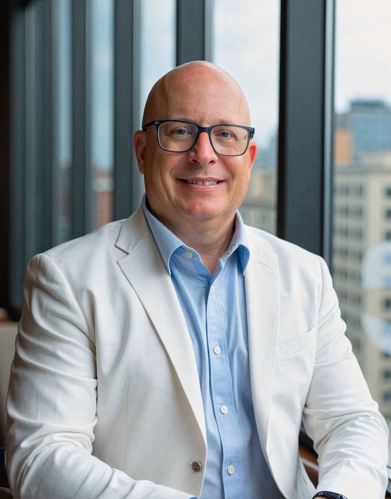 Man in a light-colored suit and glasses smiles, sitting near a window with a city view.