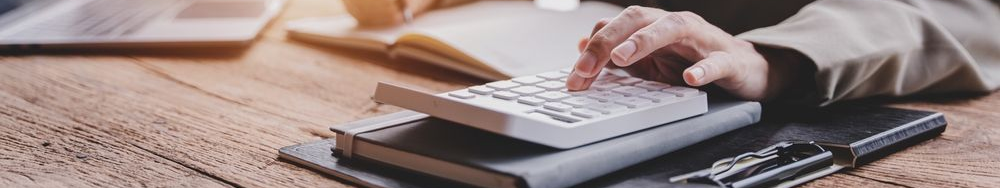 Person's hand using a calculator on a desk with a notebook, pen, and laptop.