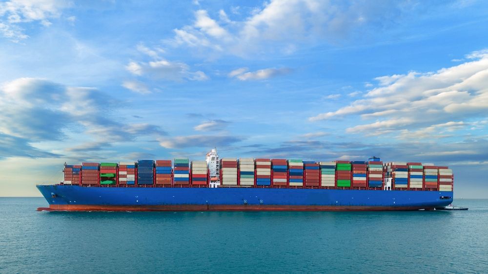 A large blue cargo ship fully loaded with colorful containers sailing on a calm ocean under a blue, cloudy sky.