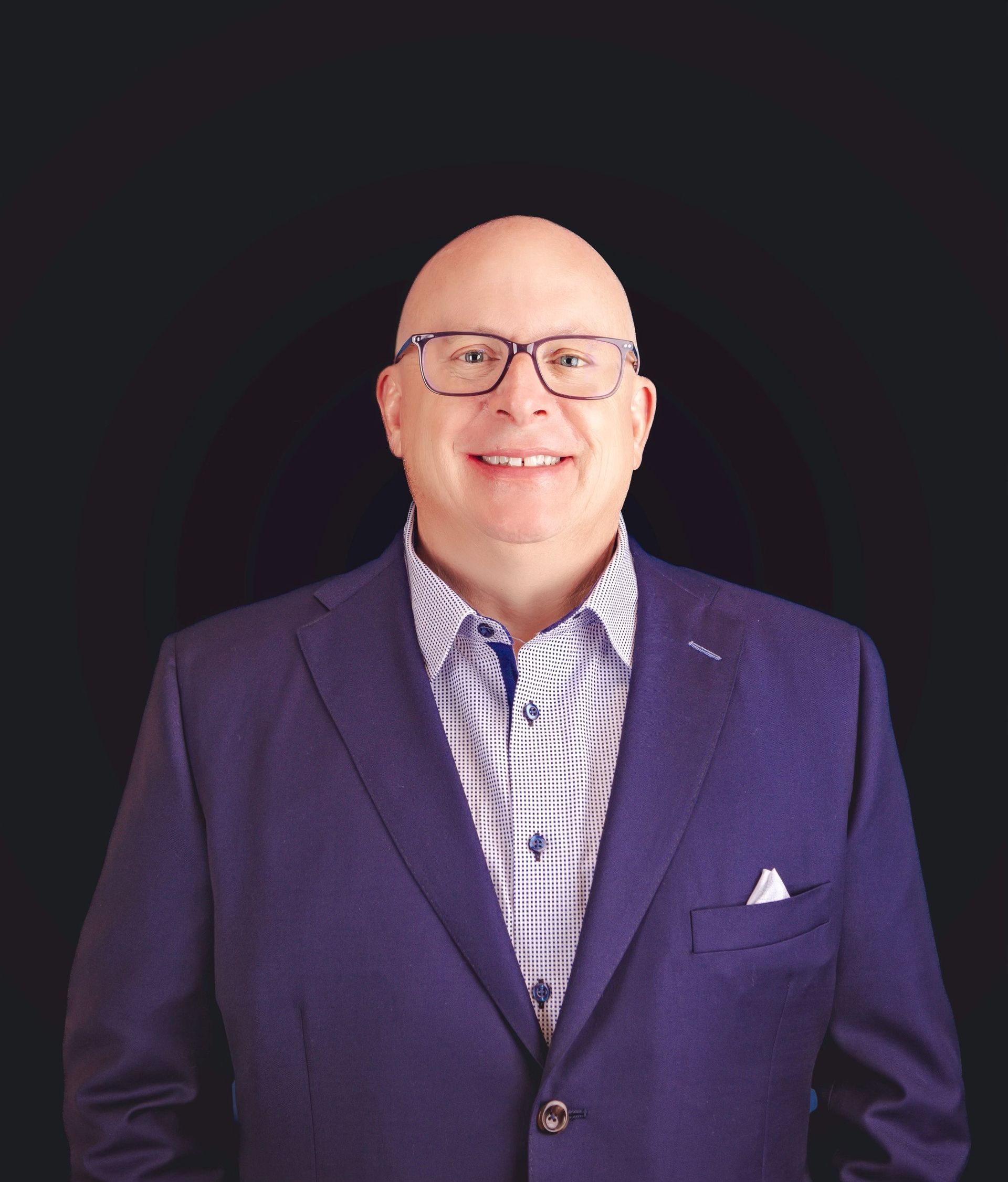 A bald man wearing glasses and a purple suit is smiling for the camera.
