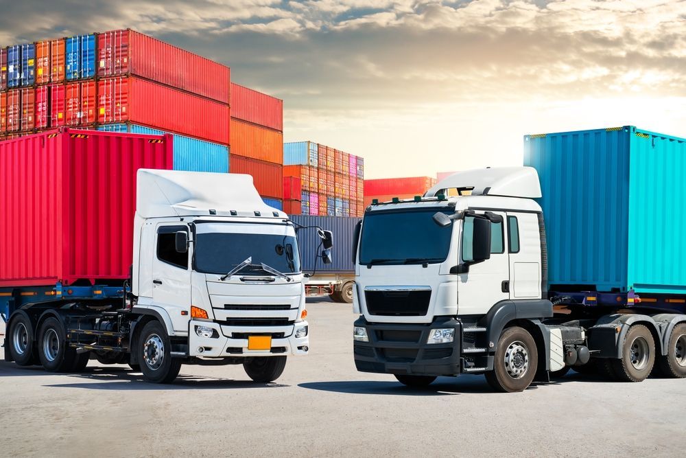 Two semi-trucks with shipping containers in a cargo yard, colorful containers stacked in the background, sunset.