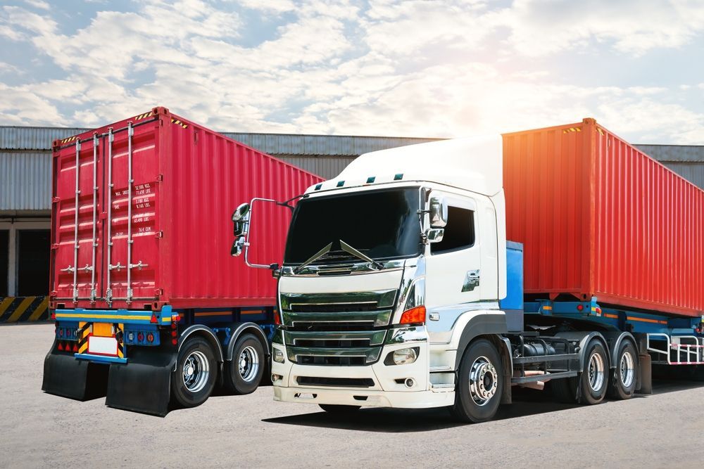 Two semi-trucks with red cargo containers parked on a paved lot; warehouse in the background.