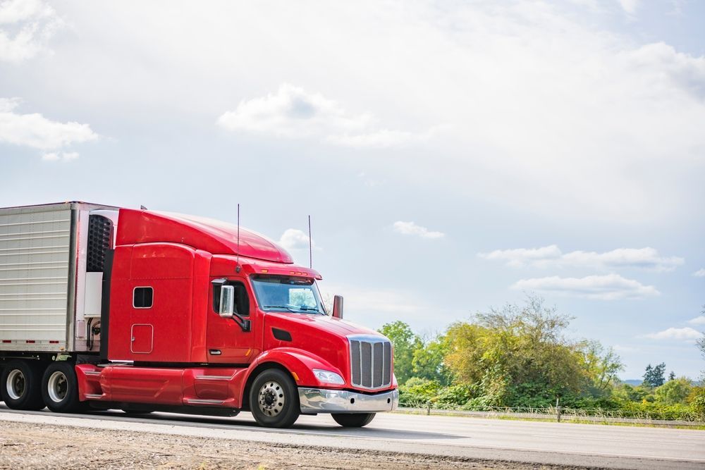 Red semi-truck parked on a roadside under a bright cloudy sky.