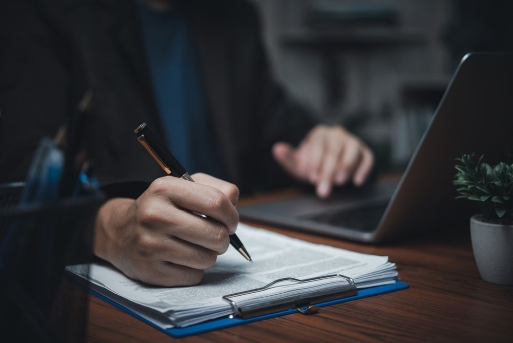 A man is writing on a clipboard while using a laptop computer.