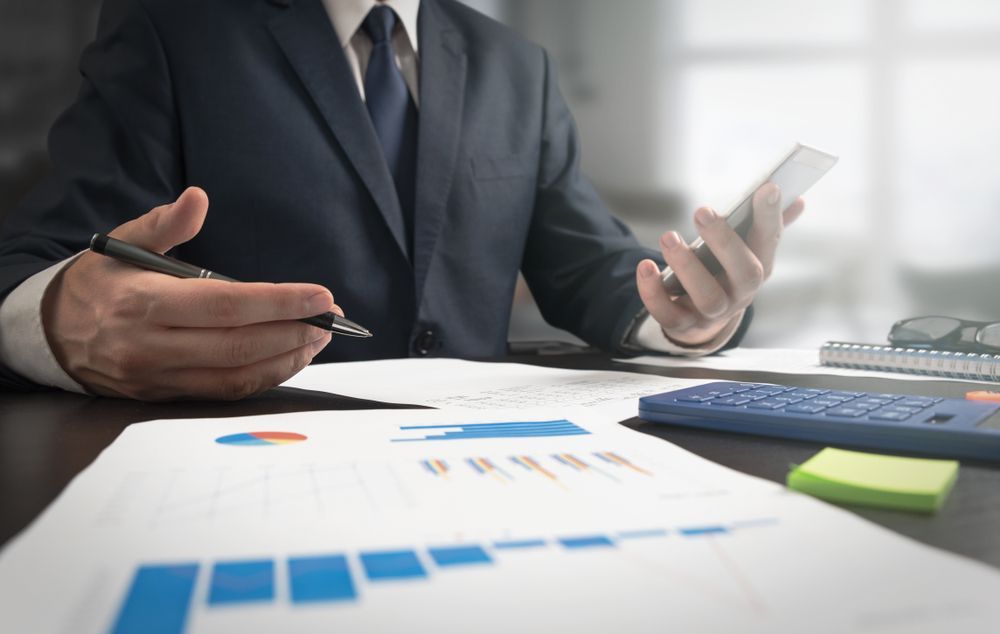 Person in suit reviewing financial charts, using a phone and pen at a desk.