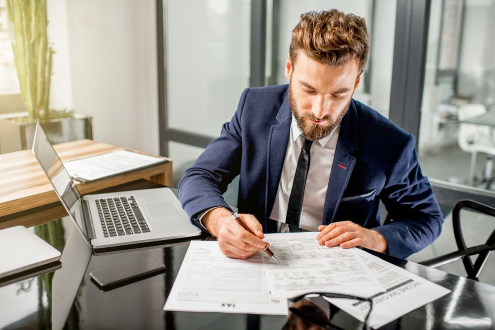 Man in suit reviewing paperwork at a desk with a laptop and eyeglasses.