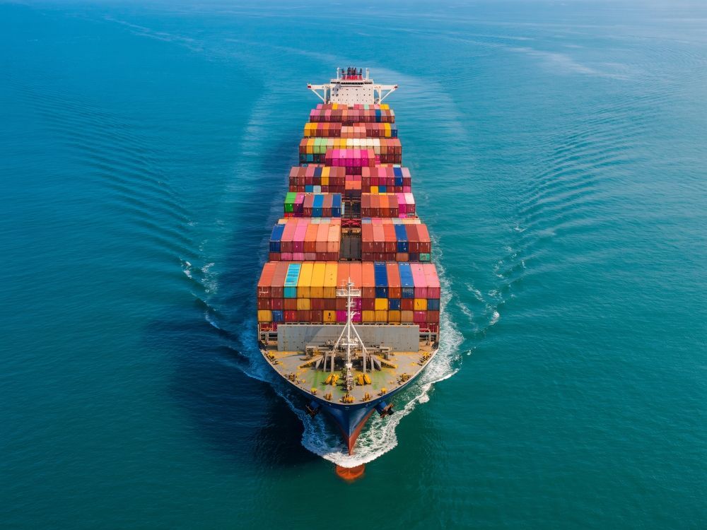 A large container ship loaded with colorful shipping crates travels through blue, open water, creating a white wake.