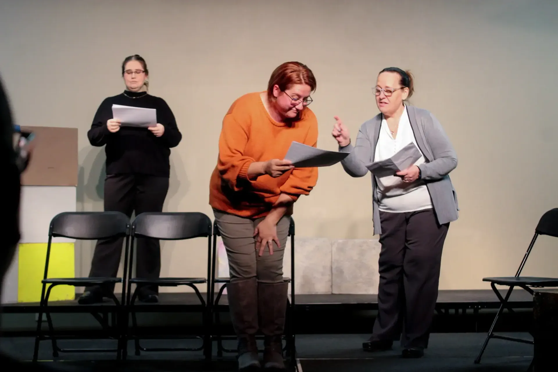 Three people on stage reading papers. One points, one bows, another stands still.