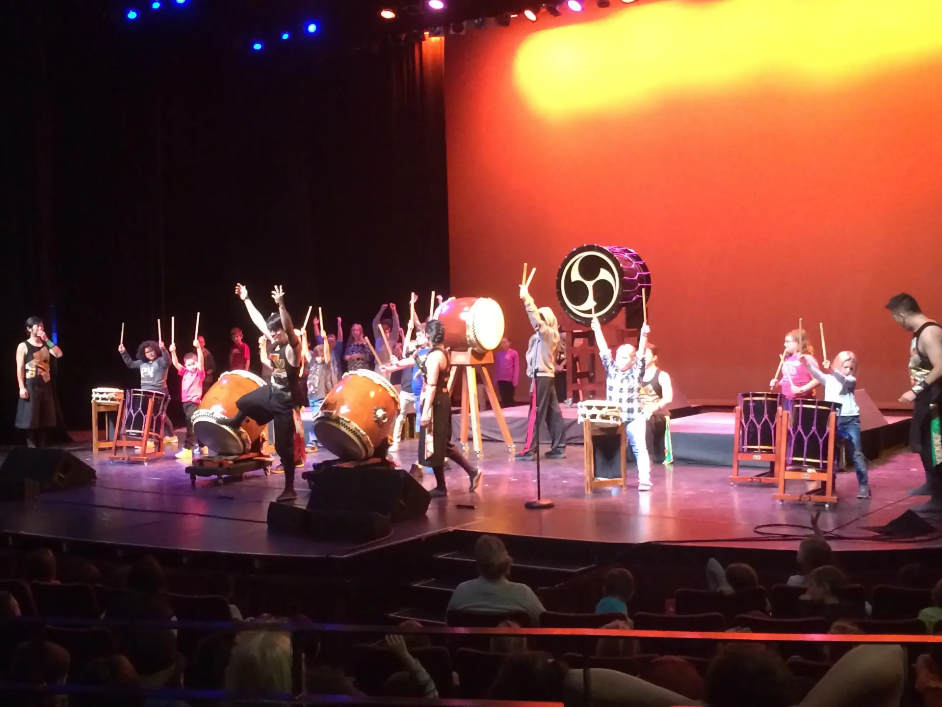Taiko drummers perform on stage with raised arms, instruments lit by orange backdrop.