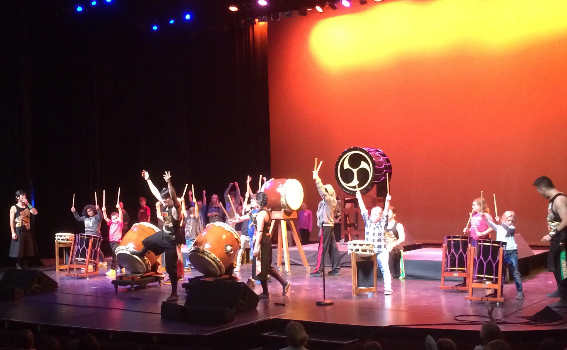 Taiko drummers on stage, performing with drums and mallets, against an orange backdrop.