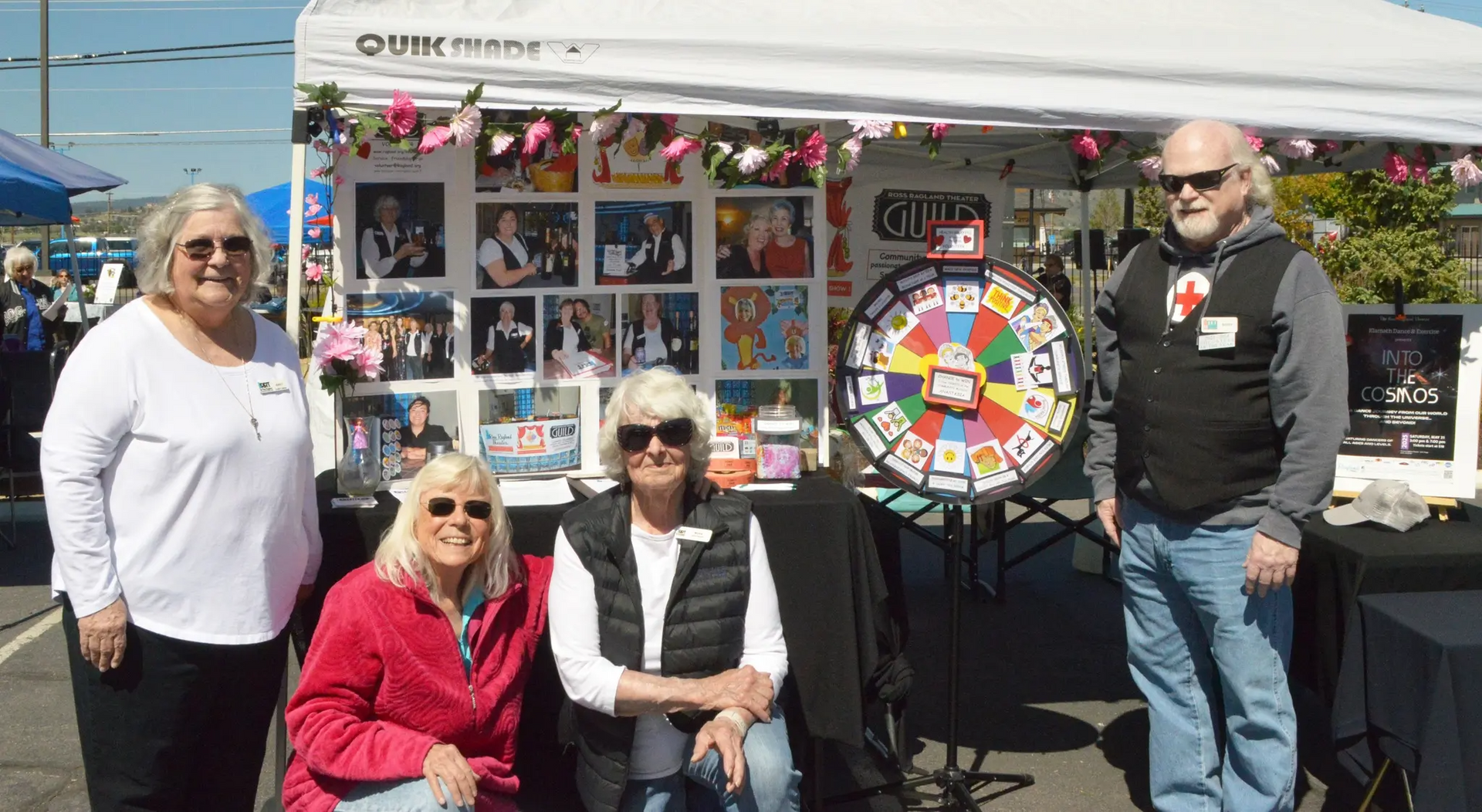 Four people at a booth decorated with photos and a prize wheel. They are outdoors under a tent.