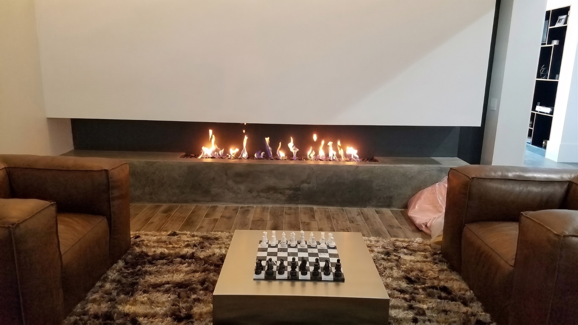 Modern lounge with a long fireplace, two brown armchairs, and a chessboard on a coffee table.