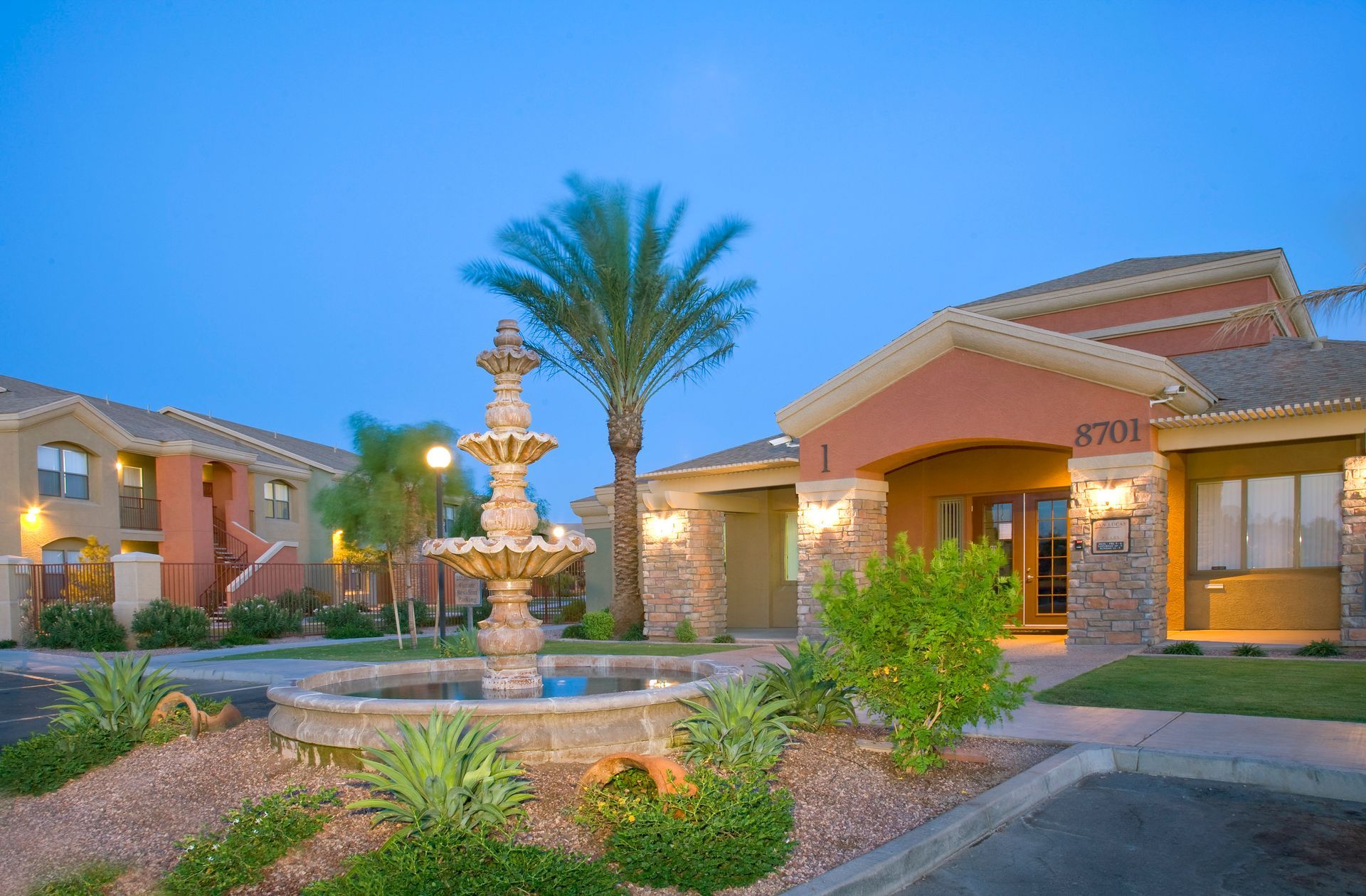 A three-tiered fountain stands in front of a tan apartment complex at twilight with a palm tree and landscaping.