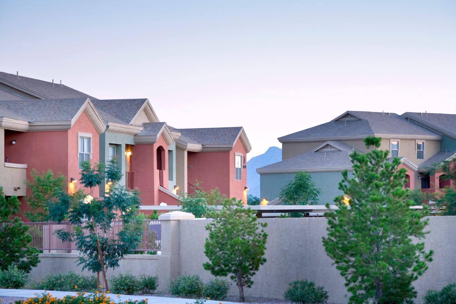 Multi-story apartment buildings with stucco walls in earth tones stand behind a decorative stone wall and small trees.