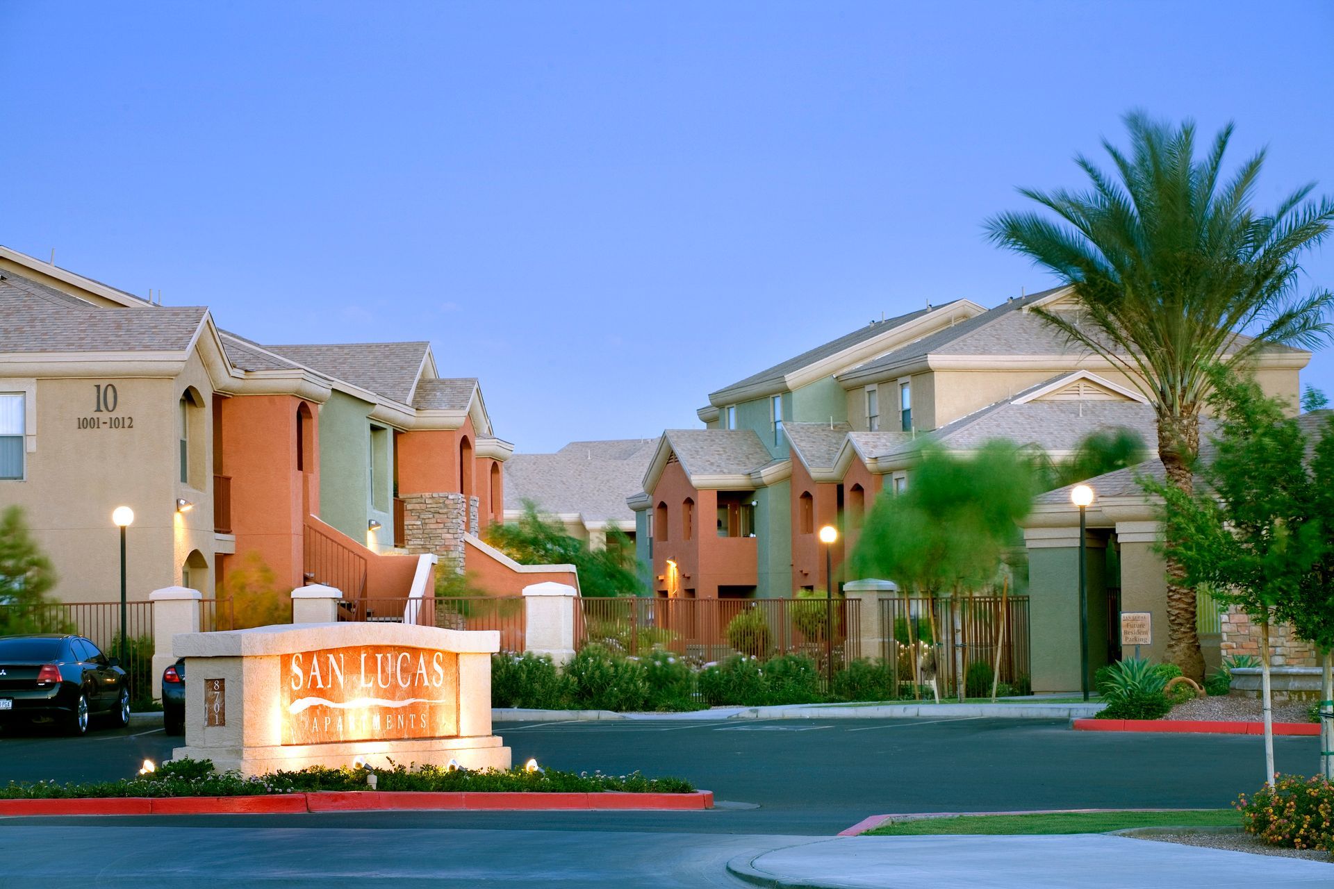 The San Lucas apartment complex entrance at dusk, featuring tan and orange buildings, a palm tree, and a stone sign.