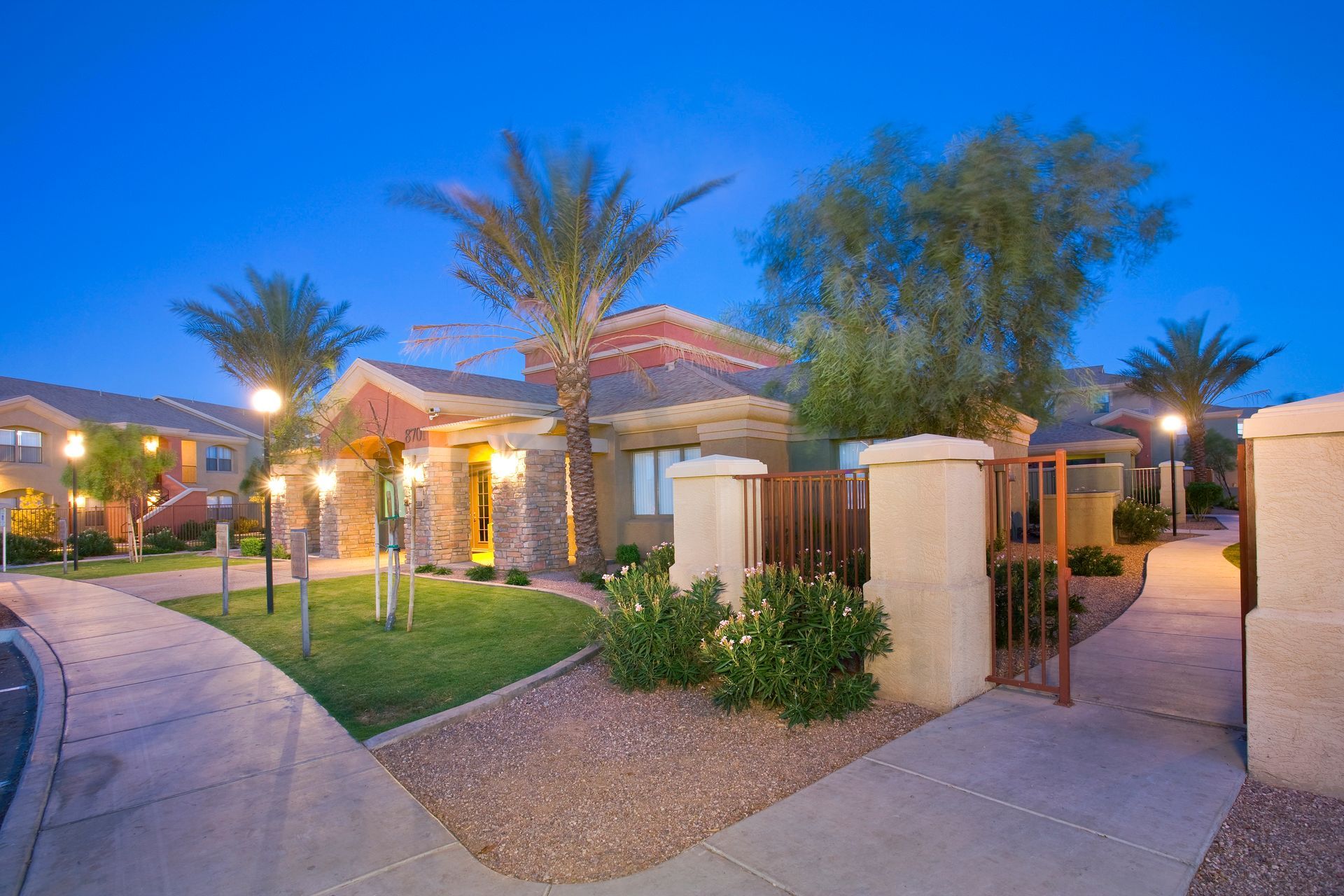 An apartment complex entrance with palm trees, landscaped grass, and glowing streetlights at twilight.
