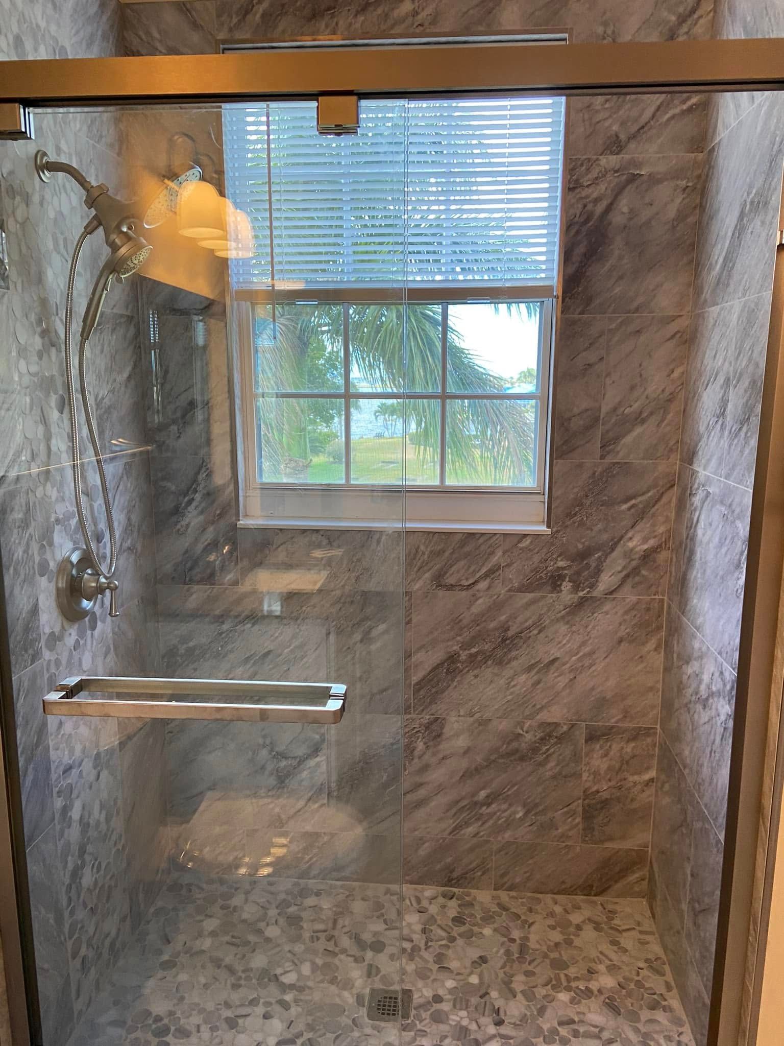 Stone-tiled walk-in shower with glass door, built-in shelf, and window with blinds.