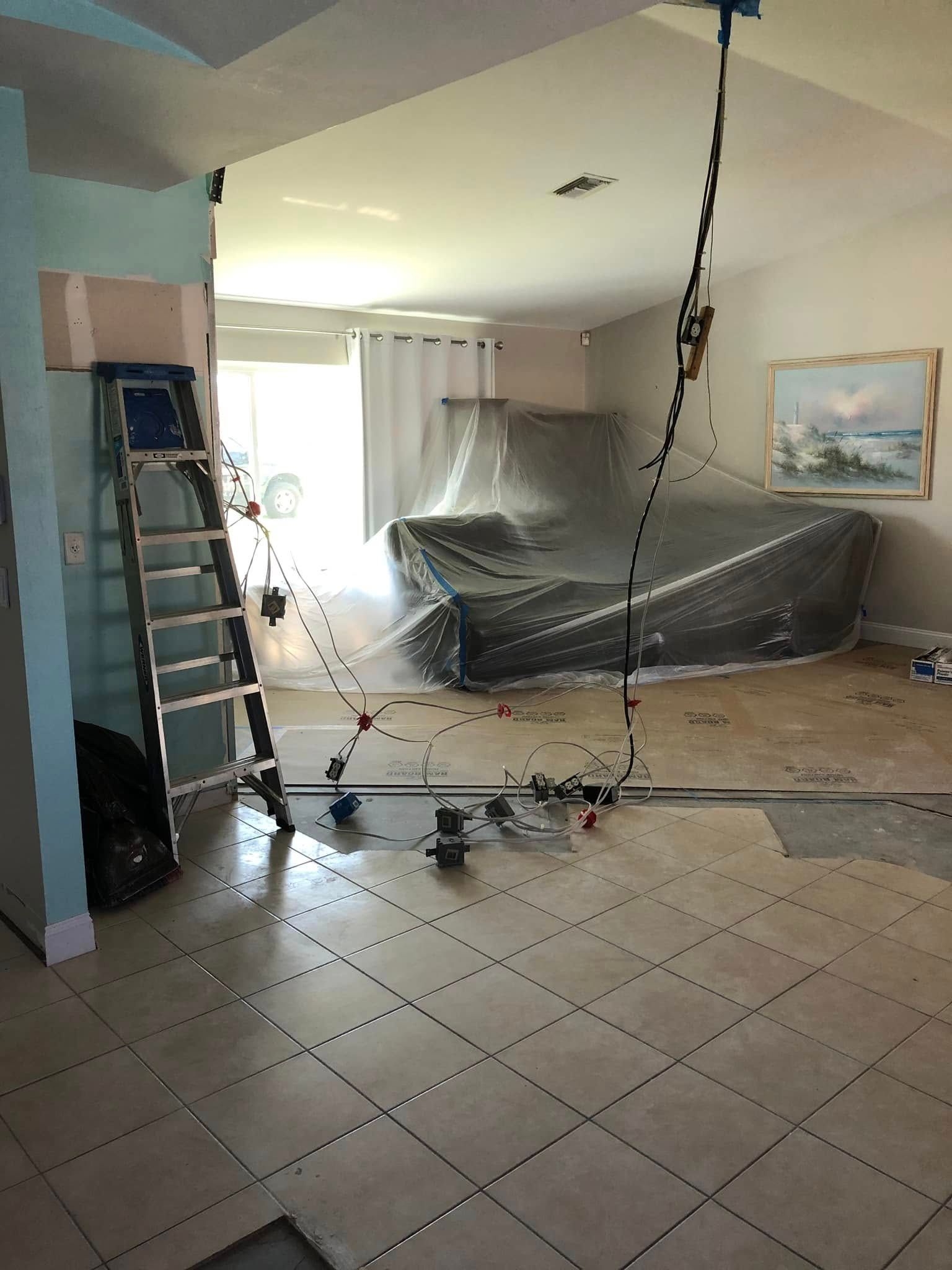 Living room under renovation, furniture wrapped in plastic, ladder and hanging wires visible