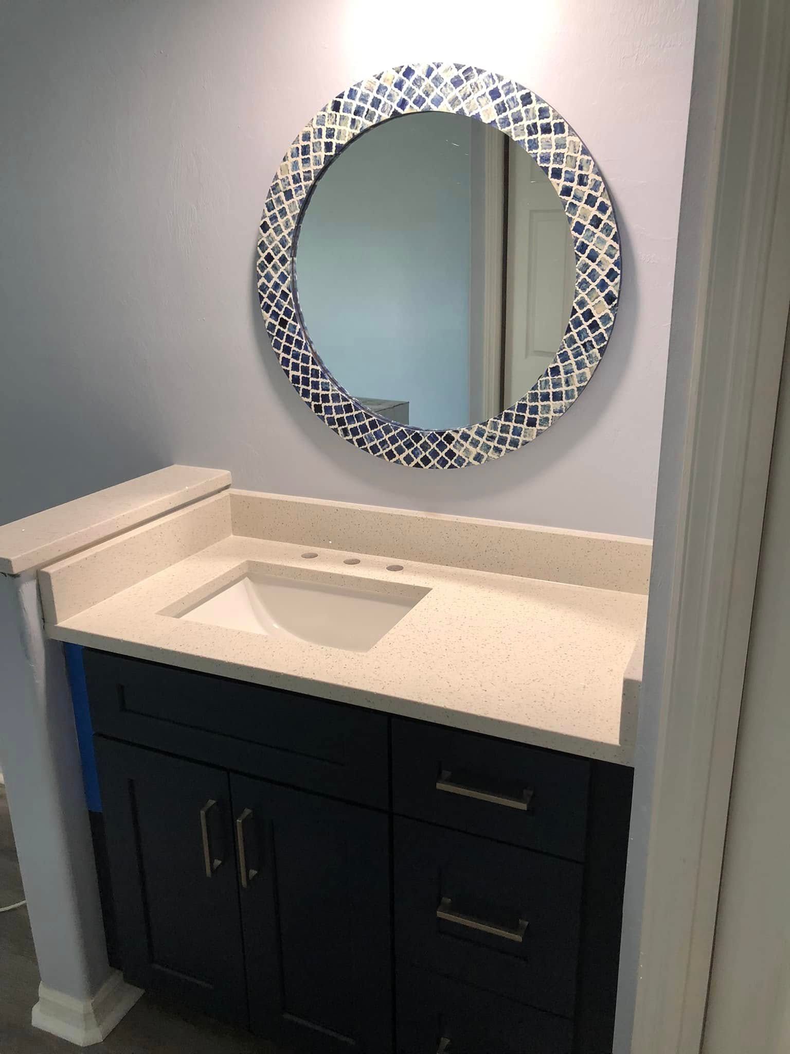 Bathroom vanity with round mosaic mirror, white sink, and dark cabinet against a light wall