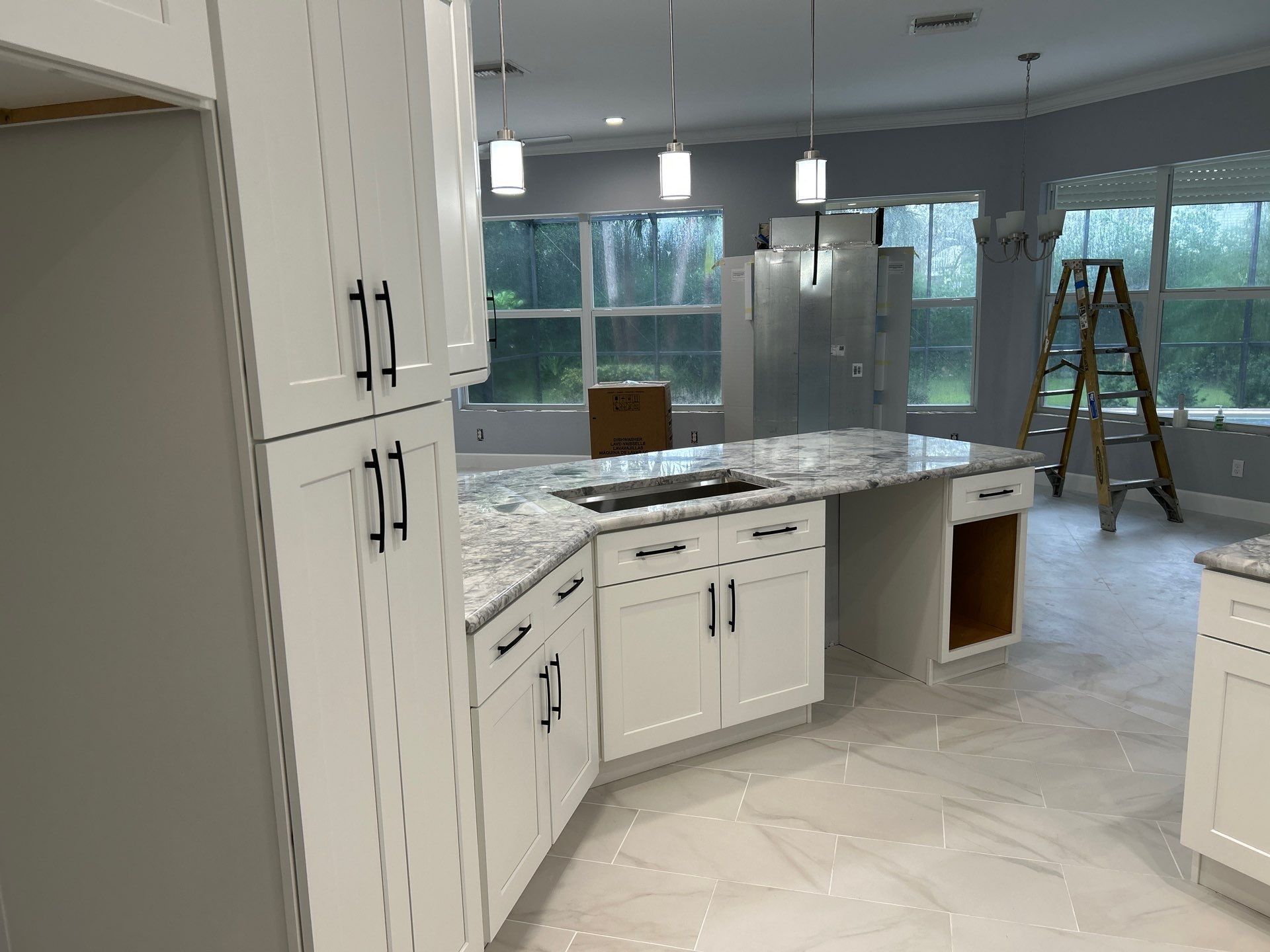 Bright kitchen with white cabinets, marble island, stainless fridge, and a ladder in the background.