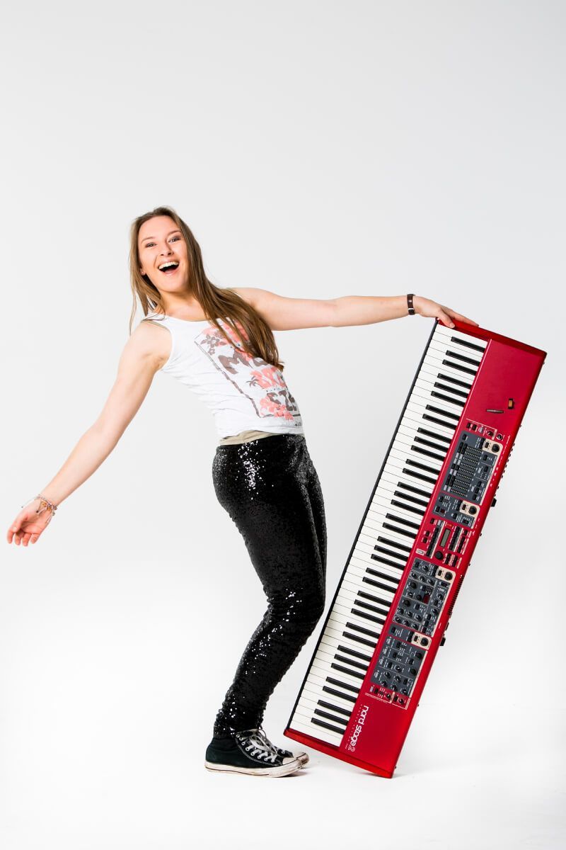 Pianodocent Charlotte Hebels uit Amersfoort lacht naast een rood toetsenbord en leunt er tegenaan, tegen een witte achtergrond.