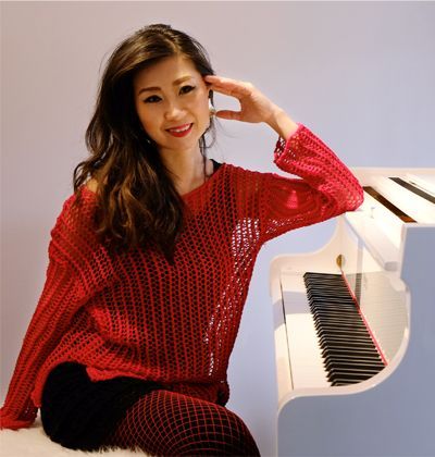 Pianodocent Cindy Joe in een rode trui zit naast een witte piano, glimlachend met haar hand bij haar gezicht.