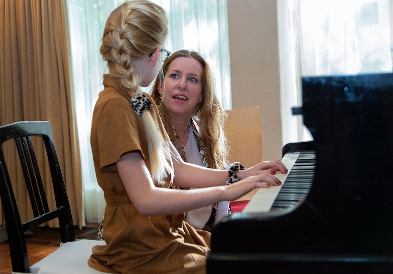 The piano Academy muziekdocent en leerling zitten aan een piano, de ene speelt terwijl de andere toekijkt in een lichte kamer.