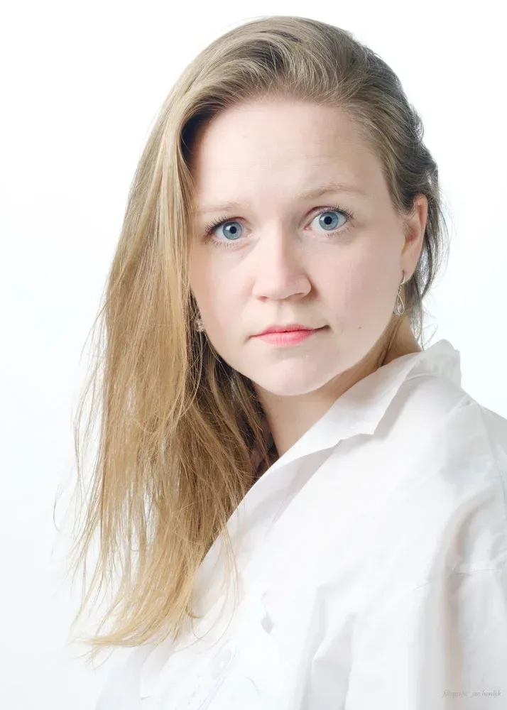 Pianodocent Ellyne Wieringa uit Rotterdam met lang blond haar in een witte blouse, die tegen een witte achtergrond in de camera kijkt.