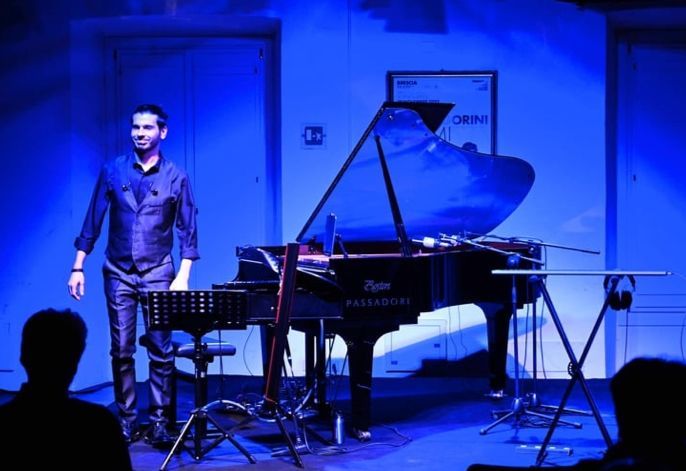Pianodocent Paolo Gorini uit Amsterdam staat naast een vleugel op een blauwverlicht podium, met silhouetten van het publiek op de voorgrond.