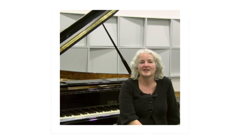 Pianodocent Juliette van Capelleveen zit naast een vleugel in een lichte kamer met witte planken.