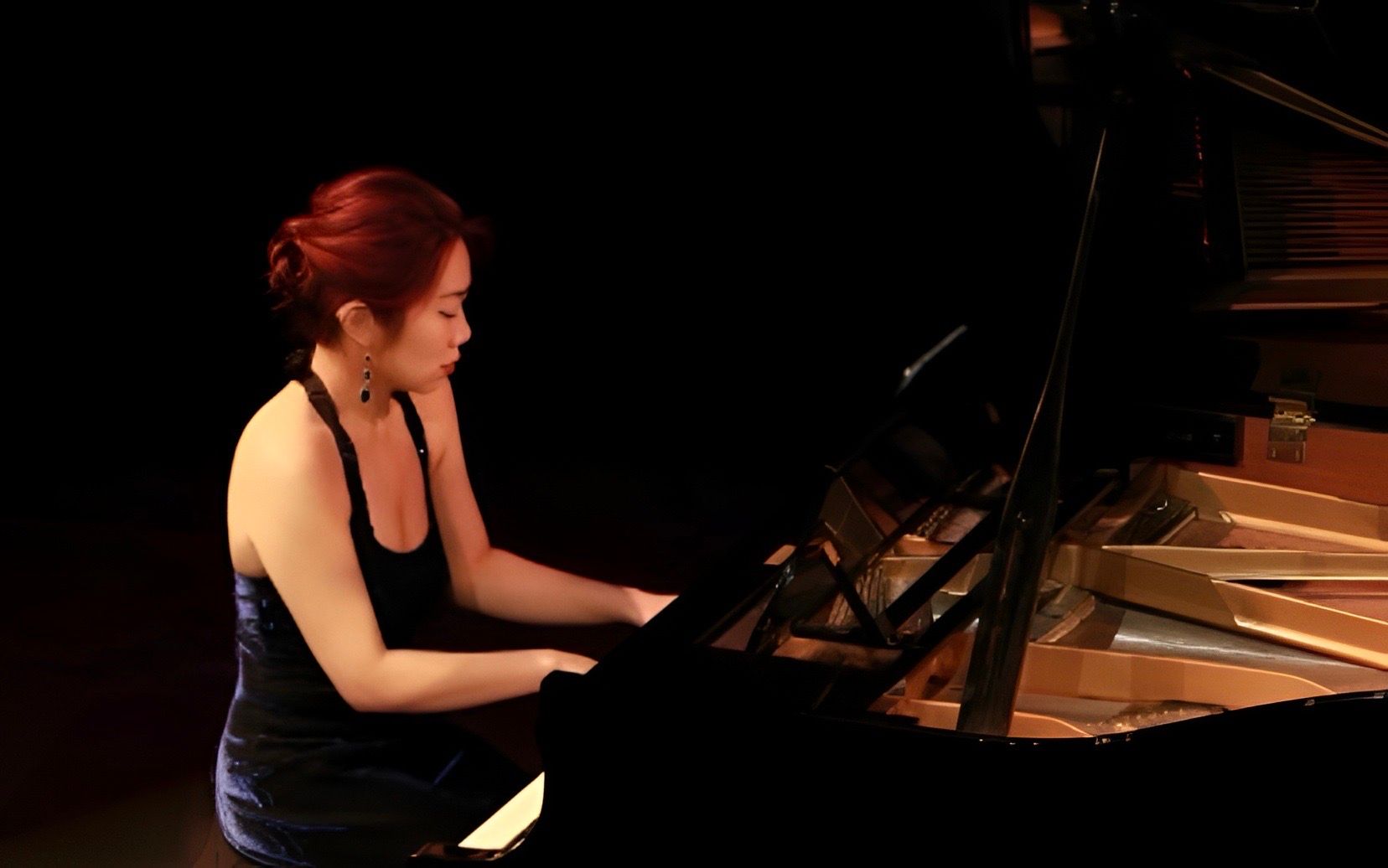 Pianodocent Yuqi Han uit Utrecht in een zwarte jurk speelt piano op een vleugel op een podium onder warm licht.