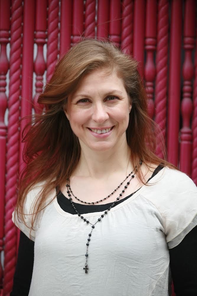 Janece Milos, pianodocent uit Amsterdamin een witte top staat voor een rode, gestructureerde achtergrond.
