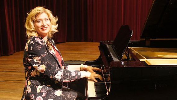 Kristina Appel, pianodocent uit Zaandam en Oostzaan speelt op een zwarte vleugel op het podium en lacht naast rode gordijnen.